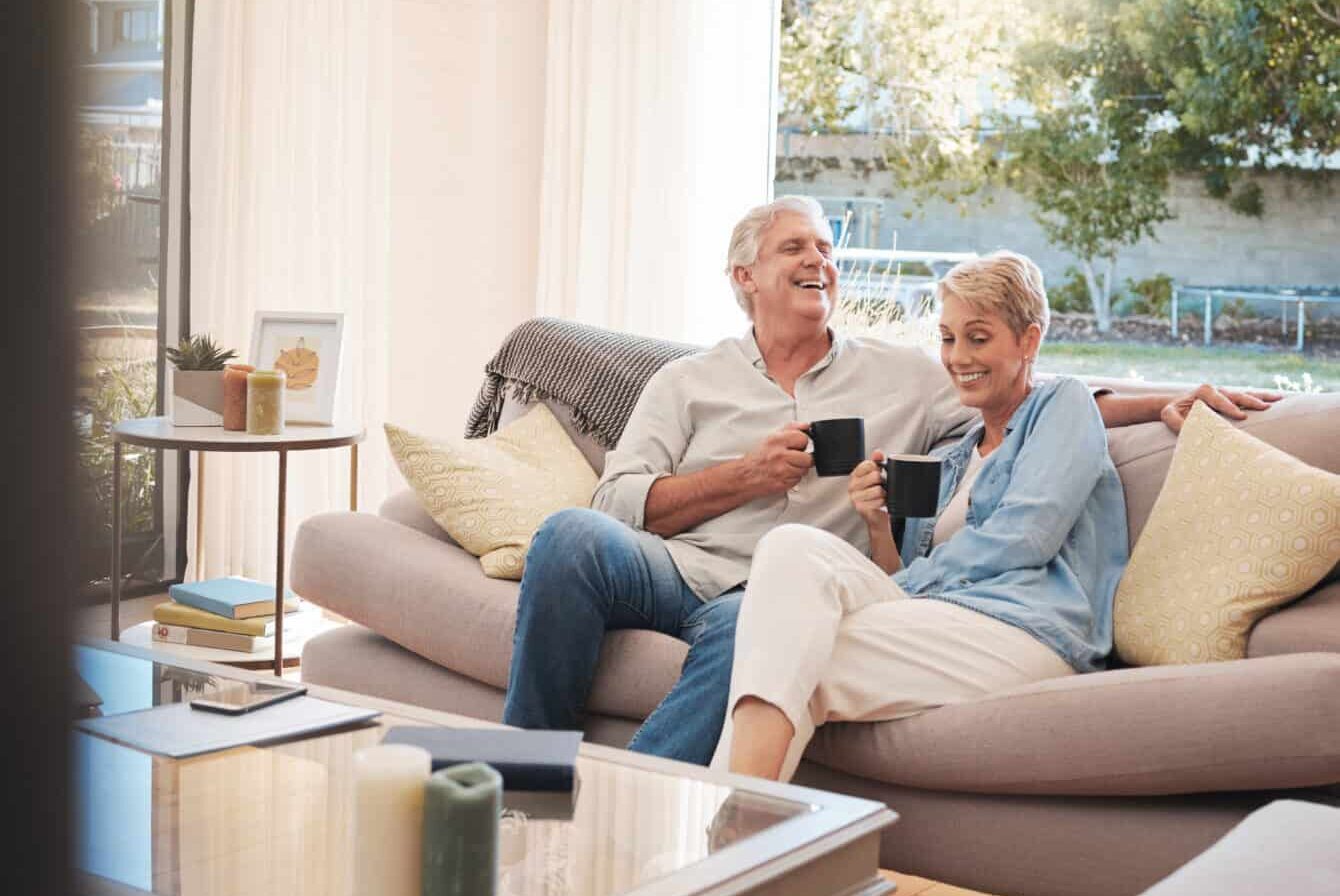 Senior couple with coffee or tea and relax on sofa in their living room happy with retirement, real estate and lifestyle. Elderly people drink on couch or in lounge together while talking of marriage.